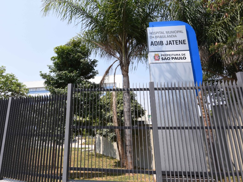 Fachada do Hospital Municipal da Brasilândia. Em primeiro plano está o portão na cor chumbo e ao fundo o hospital com algumas árvores. Do lado direito da foto há um totem onde está escrito Hospital Municipal da Brasilândia Adib Jatene e abaixo Prefeitura de São Paulo